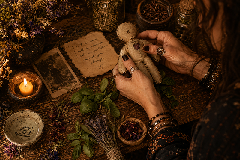 A person holds a small handmade cloth doll with a stitched heart on its chest over a rustic wooden table, surrounded by a lit candle, dried flowers and herbs, fresh greenery, small bowls and jars, an old photograph, and a handwritten note about courage, protection, and love.